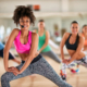 A diverse group of people performing a dance exercise stretching routine in a gym. Dark skinned woman at the front in a pink tank top and black leggings wears a microphone and instructs the class.