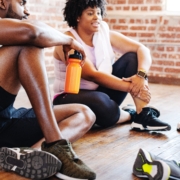 Group of African American men and women in athletic wear, resting after a workout. The man is holding an orange water bottle. They are sitting on a wooden floor. Exercise for better health and wellness. Diverse group workout at fitness.