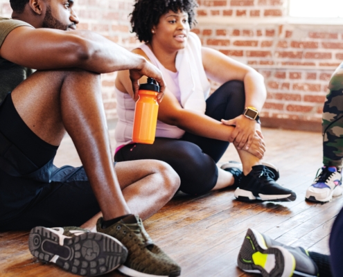 Group of African American men and women in athletic wear, resting after a workout. The man is holding an orange water bottle. They are sitting on a wooden floor. Exercise for better health and wellness. Diverse group workout at fitness.