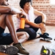 Group of African American men and women in athletic wear, resting after a workout. The man is holding an orange water bottle. They are sitting on a wooden floor. Exercise for better health and wellness. Diverse group workout at fitness.