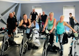 Mixed age and gender group of people dressed in gym clothing, standing next to spinning bikes.