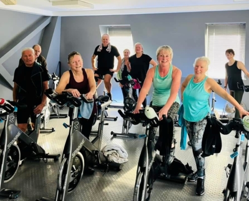 Mixed age and gender group of people dressed in gym clothing, standing next to spinning bikes.