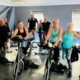 Mixed age and gender group of people dressed in gym clothing, standing next to spinning bikes.