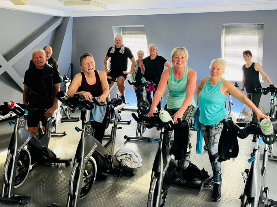 Mixed age and gender group of people dressed in gym clothing, standing next to spinning bikes.