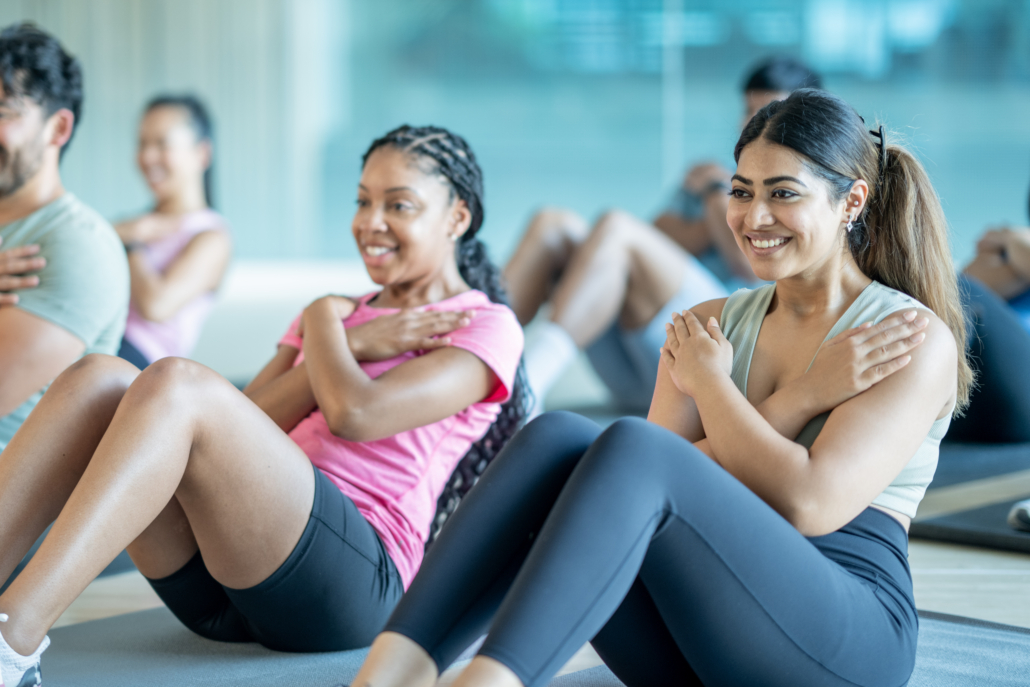 A small group of young adults sit spread apart on yoga mats in a gym, as they participate in a fitness class together. They are each dressed comfortably and are working through a series of sit-up repetitions.