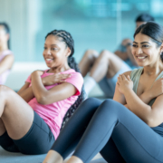 A small group of young adults sit spread apart on yoga mats in a gym, as they participate in a fitness class together. They are each dressed comfortably and are working through a series of sit-up repetitions.