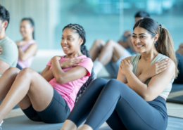 A small group of young adults sit spread apart on yoga mats in a gym, as they participate in a fitness class together. They are each dressed comfortably and are working through a series of sit-up repetitions.