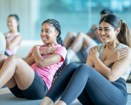 A small group of young adults sit spread apart on yoga mats in a gym, as they participate in a fitness class together. They are each dressed comfortably and are working through a series of sit-up repetitions.