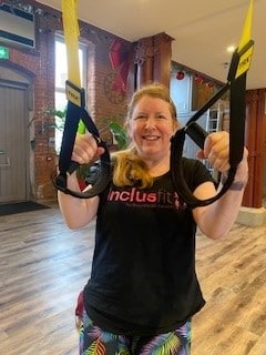 Sam James (white woman with brown hair in a ponytail) in a black tshirt with Inclus on the front, brases on some exercise equipment.