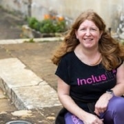 Sam James (white, long brown hair) sits in a black Inclusfit tshirt and jeans in a garden.