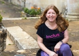 Sam James (white, long brown hair) sits in a black Inclusfit tshirt and jeans in a garden.