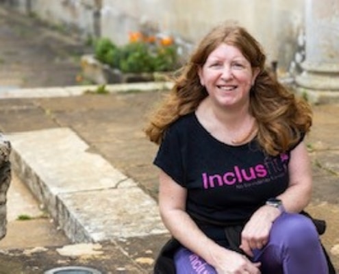 Sam James (white, long brown hair) sits in a black Inclusfit tshirt and jeans in a garden.