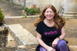 Sam James (white, long brown hair) sits in a black Inclusfit tshirt and jeans in a garden.