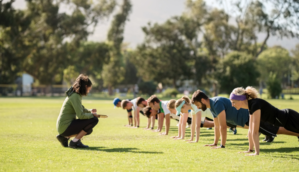 Group of diverse adults in exercise clothing doing planks in an open grass space with an instructor crouched down in front of them, coaching them.