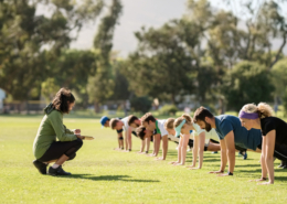 Group of diverse adults in exercise clothing doing planks in an open grass space with an instructor crouched down in front of them, coaching them.
