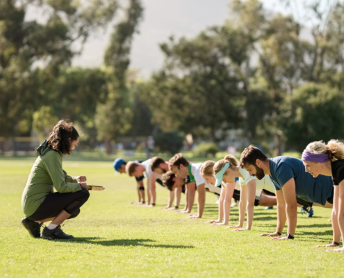 Group of diverse adults in exercise clothing doing planks in an open grass space with an instructor crouched down in front of them, coaching them.
