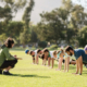 Group of diverse adults in exercise clothing doing planks in an open grass space with an instructor crouched down in front of them, coaching them.