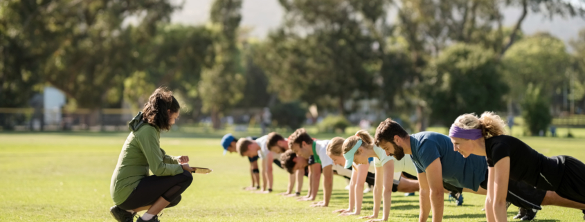 Group of diverse adults in exercise clothing doing planks in an open grass space with an instructor crouched down in front of them, coaching them.