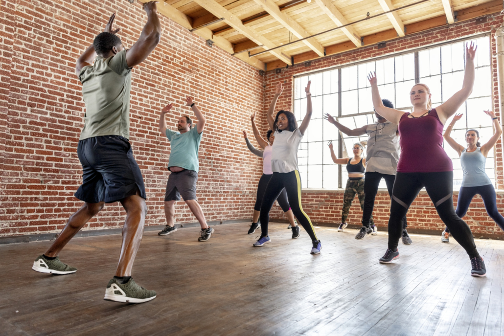 Trainer with his group fitness class doing star jumps. Fitness class is made up of diverse group of adults wearing exercise gear.